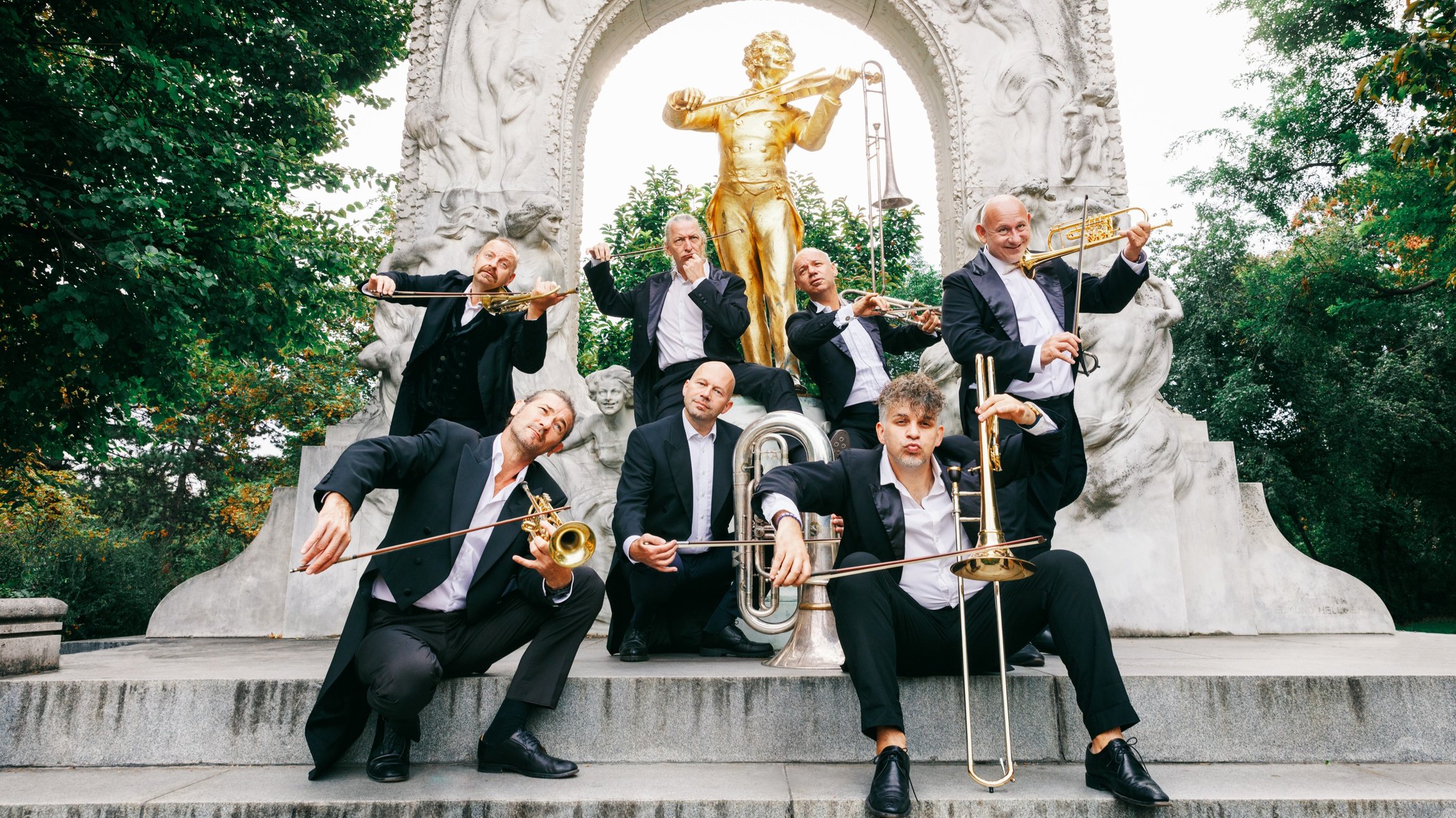 Die Mitglieder der Mnozil Brass in dunklen Anzügen mit Instrumenten auf vor einer historischen Treppe, im Hintergrund eine goldfarbene Statue