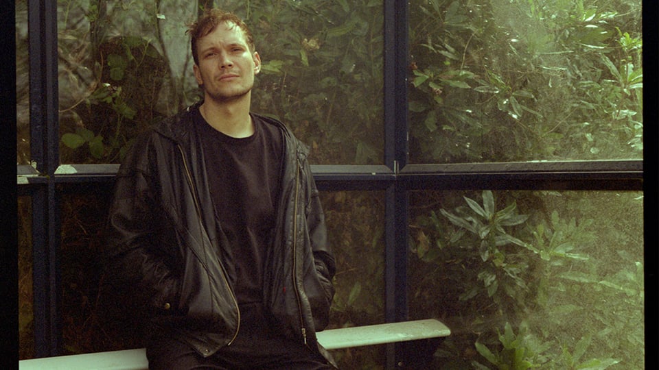 Bruno Kawelke steht vor einer Glaswand mit Pflanzen im Hintergrund, trägt eine dunkle Jacke und schaut ruhig in die Kamera, natürliche Lichtstimmung.