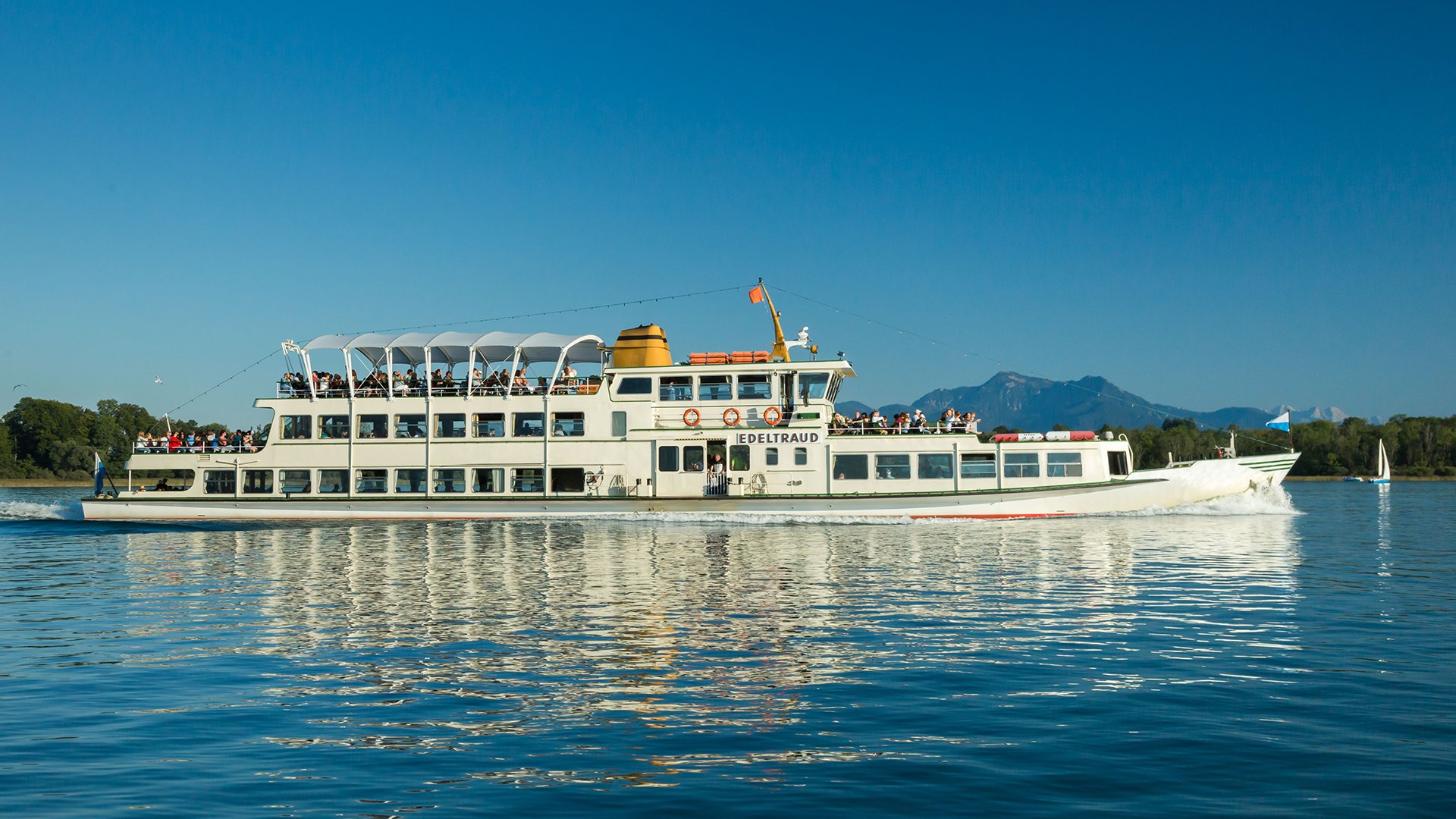 Fahrgastschiff Edeltraud mit mehreren Passagieren auf den Außendecks, unterwegs auf einem See vor Bergkulisse und blauem Himmel, mit Spiegelung des Schiffes im ruhigen Wasser