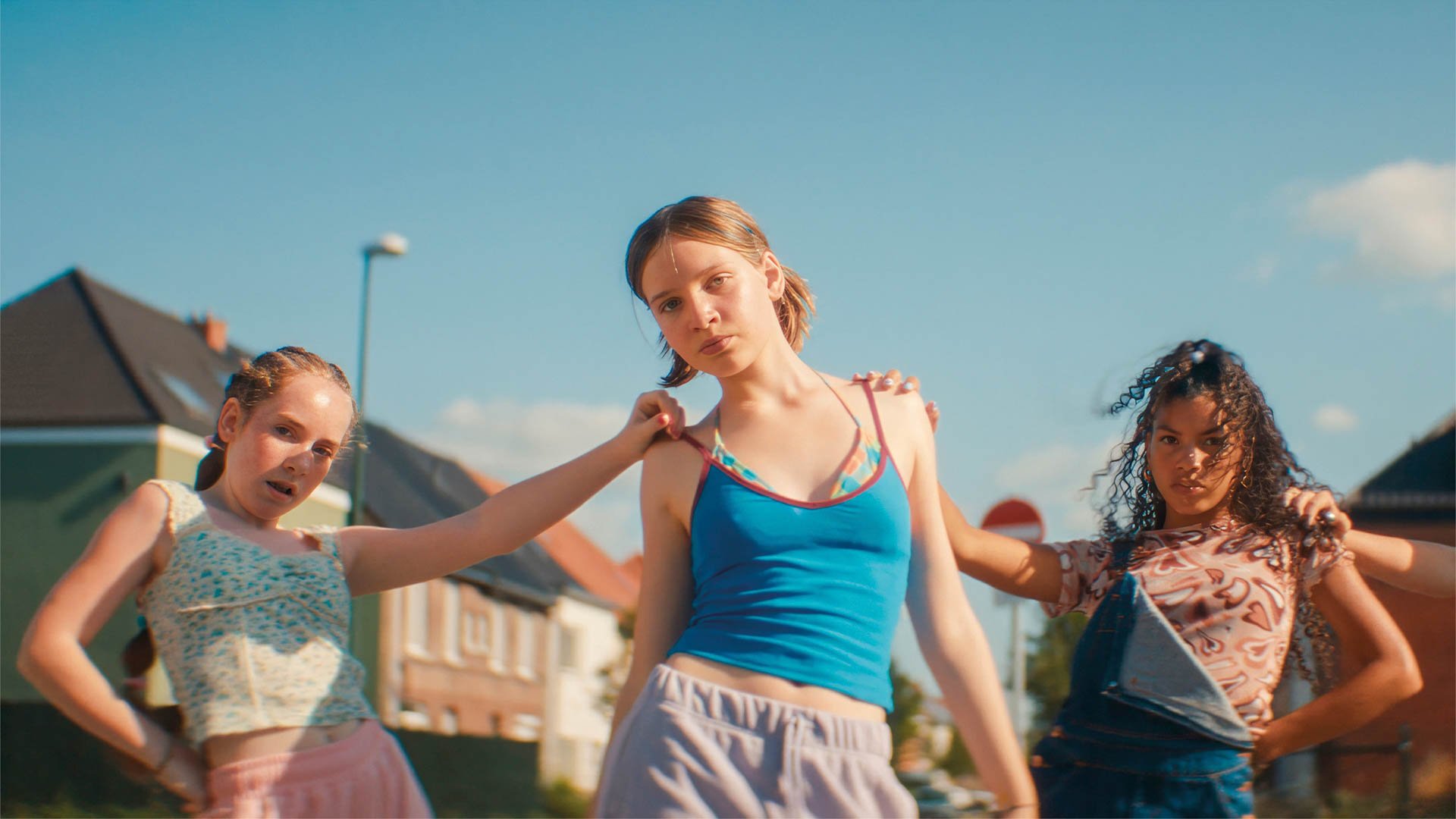Drei Kinder stehen im Freien in einer Wohngegend unter blauem Himmel; die mittlere Person trägt ein blaues Top und helle Shorts, während die beiden anderen seitlich versetzt stehen und bunte, sommerliche Kleidung tragen.