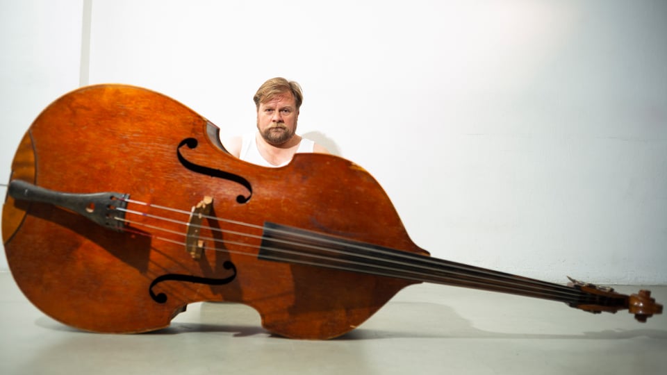 Eine Person sitzt hinter einem auf dem Boden liegenden Kontrabass vor weißem Hintergrund