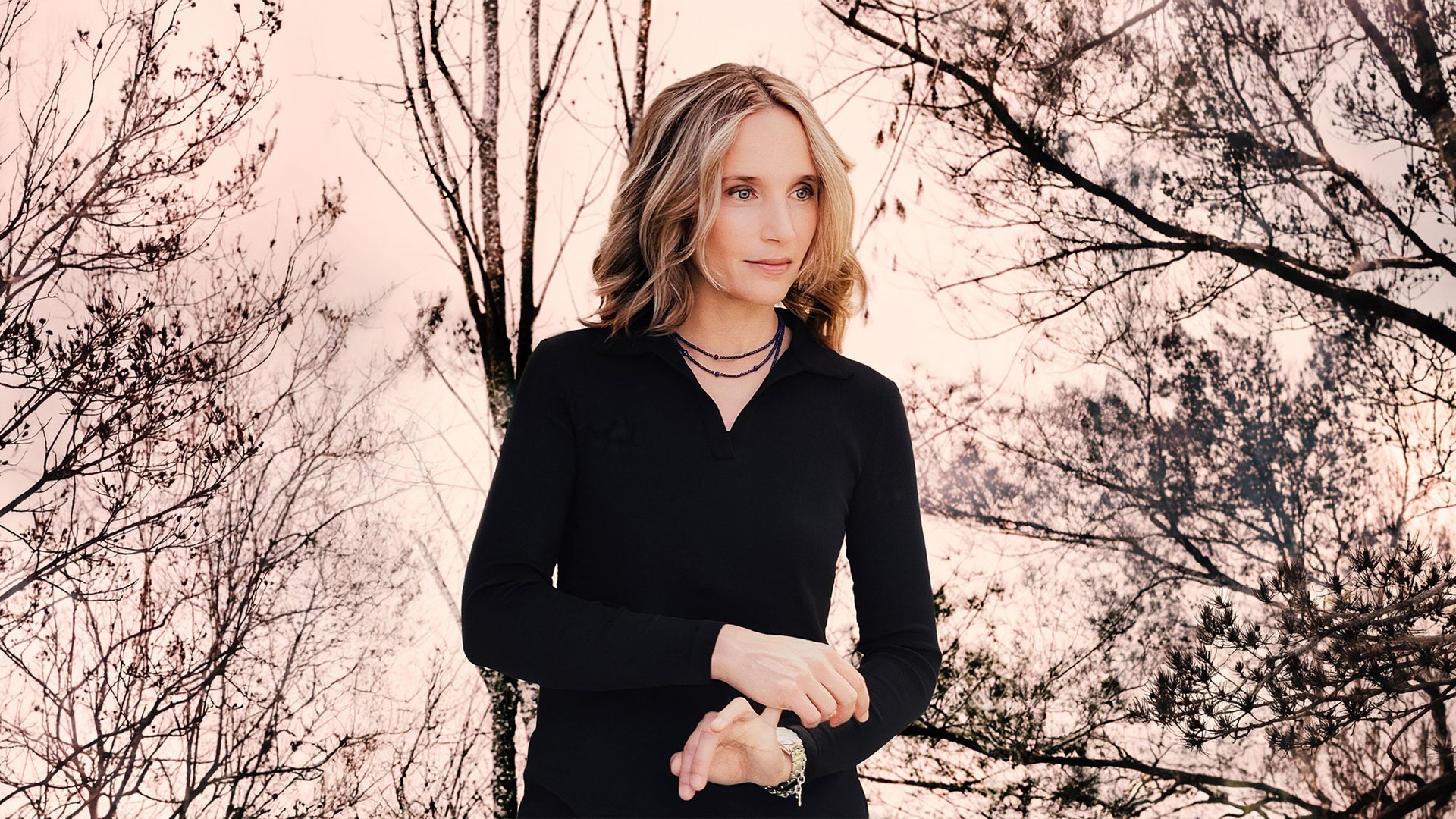 Hélène Grimaud in schwarzer Kleidung, die Hände ineinander gelegt, vor kahlen Bäumen im warmen Abendlicht