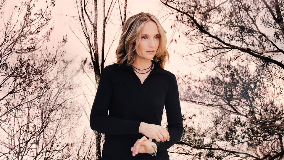 Hélène Grimaud in schwarzer Kleidung, die Hände ineinander gelegt, vor kahlen Bäumen im warmen Abendlicht
