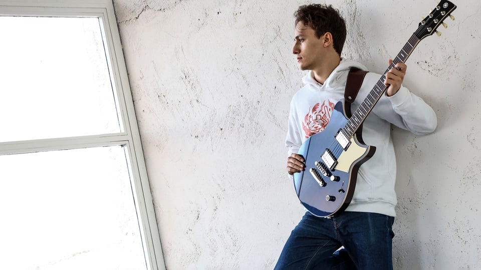 Foto für die Veranstaltung ‚MATTEO MANCUSO‘ mit dem Künstler, der sich mit einer Gitarre an eine weiße Wand lehnt.
