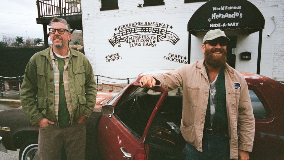 Foto für die Veranstaltung „The Black Keys“ mit den beiden Künstlern, einer von ihnen lehnt an der Tür eines bordeauxfarbenen Autos, im Hintergrund ein weißes Gebäude.