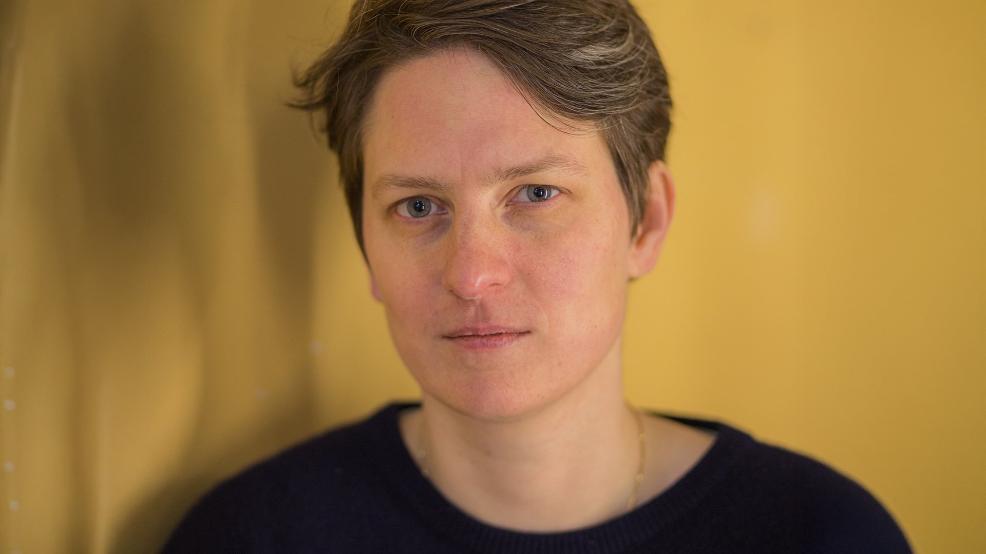 Anna Julian Mendlik in schwarzem Pullover, direkt in die Kamera blickend, vor ockergelbem verschwommenem Hintergrund