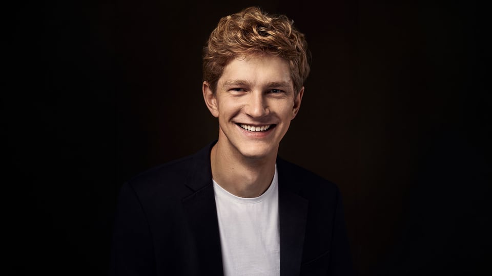 Der Künstler Jan Lisiecki in weißem Shirt und schwarzem Sakko, lachend, vor schwarzem Hintergrund 