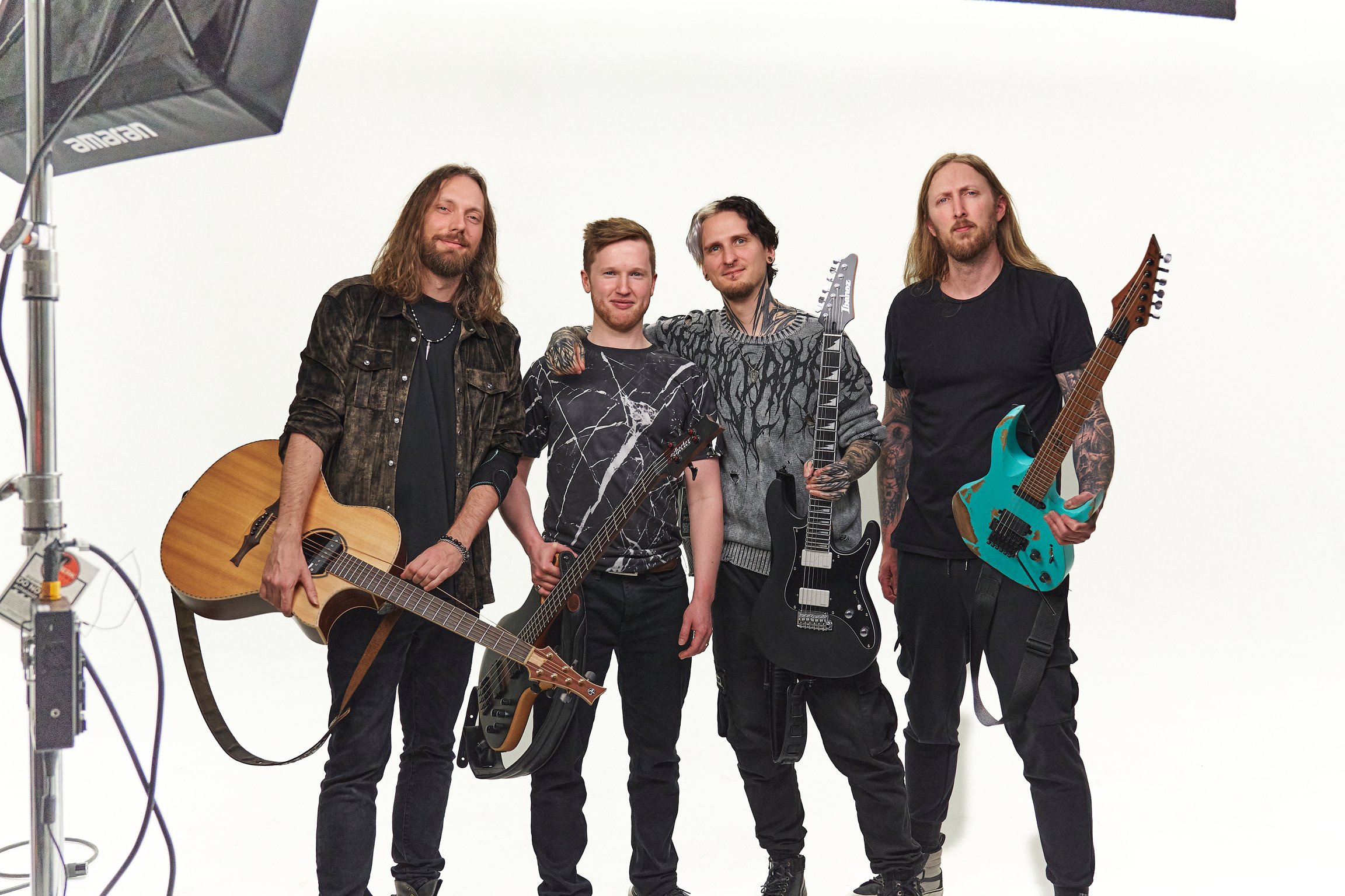 Vier Personen mit Gitarren und Bass stehen nebeneinander vor weißem Studiohintergrund; links ist ein Beleuchtungs-Setup sichtbar, die Gruppe trägt dunkle Kleidung und hält ihre Instrumente in die Kamera.