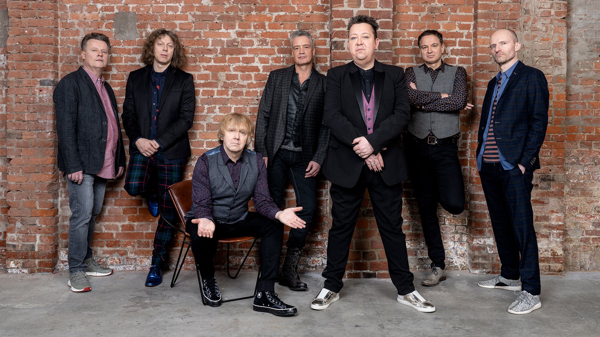 Sieben Personen der Band die Prinzen in dunkler, teils eleganter Kleidung vor roterbrauner Backsteinwand, sechs Personen stehen, eine Person sitzt auf einem Stuhl mittig vorne