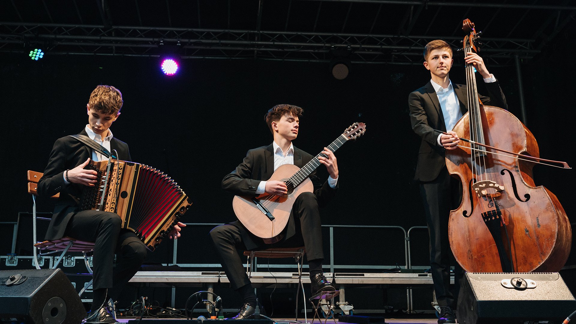 Drei junge Musiker spielen auf einer Bühne Akkordeon, Gitarre und Kontrabass.