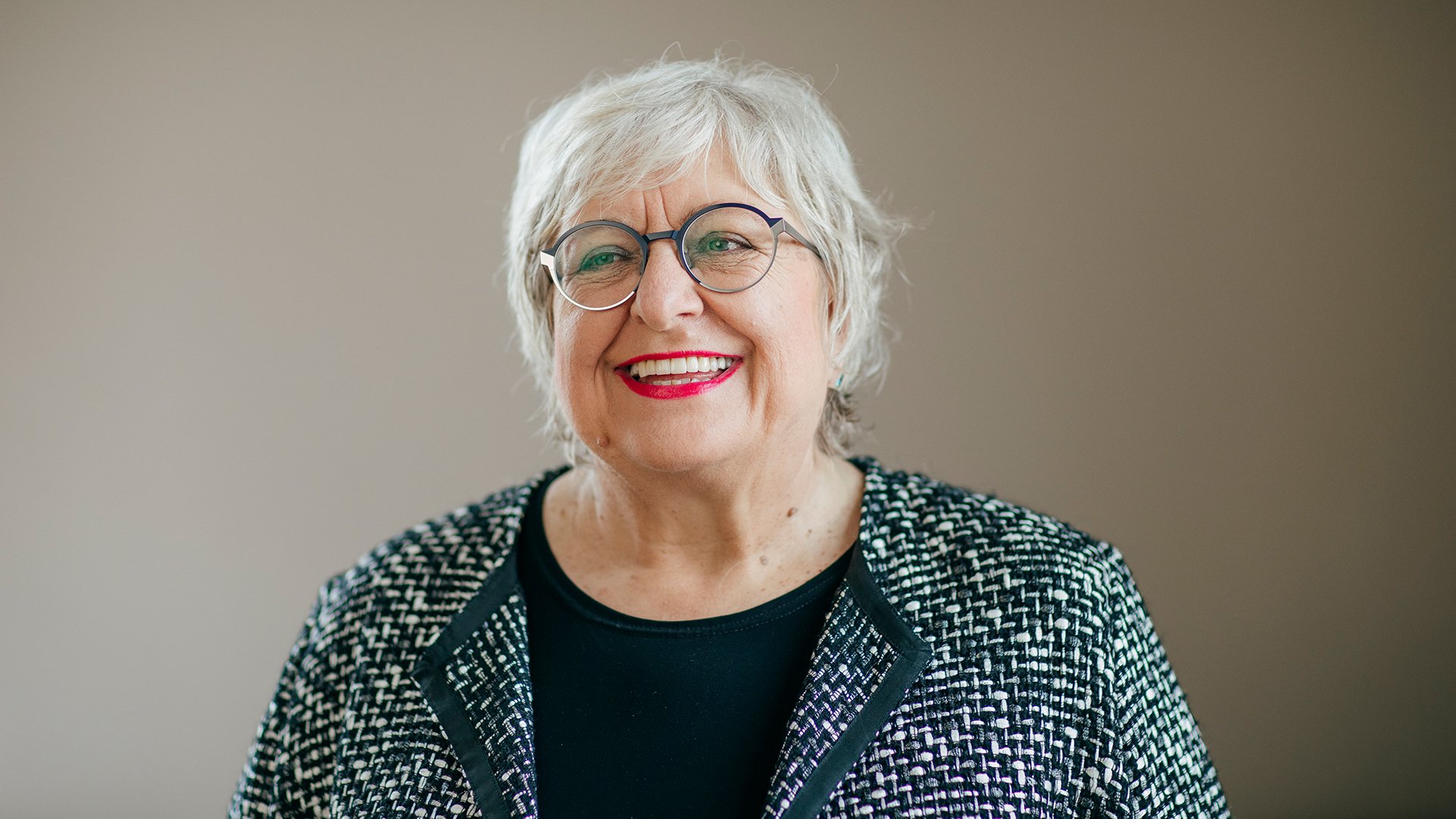 Eine lachende Dame mit weißem Haar, Brille und schwarz-weisser Jacke vor hellbraunem Hintergrund zur Veranstaltung Ladies Lunch