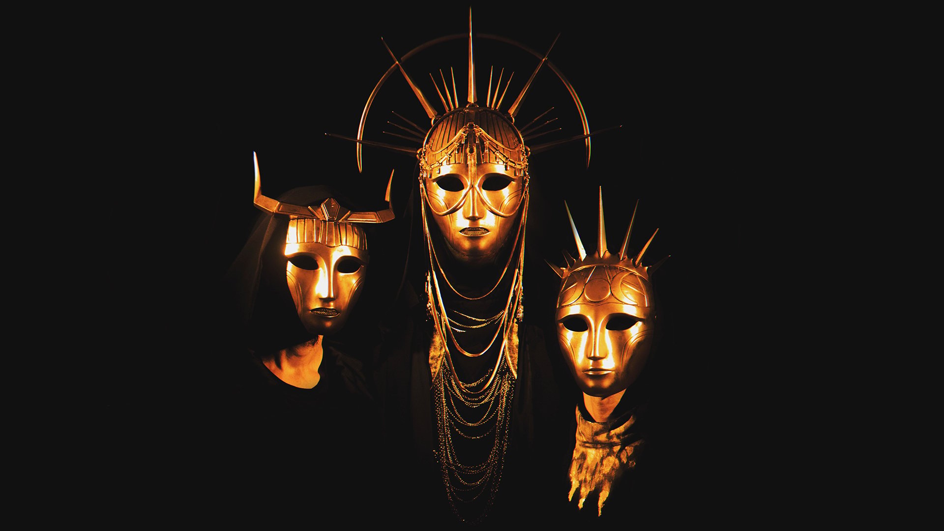 Bandfoto von Imperial Triumphant mit drei Personen in schwarzen Gewändern und goldfarbenen, kunstvoll verzierten Masken vor schwarzem Hintergrund, die mittlere Maske mit strahlenförmigem Kopfschmuck und die beiden seitlichen mit markanten Hörnern beziehungsweise Spitzen