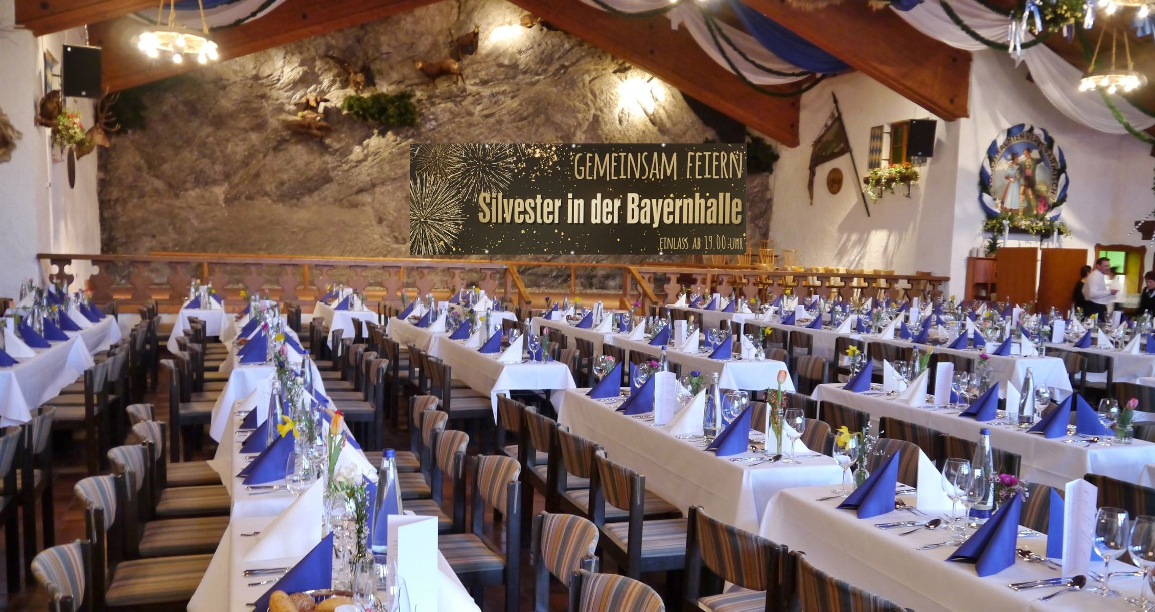 Festsaal mit weiss und blau gedeckten Tischen und Plakat im Hintergrund mit Aufschrift