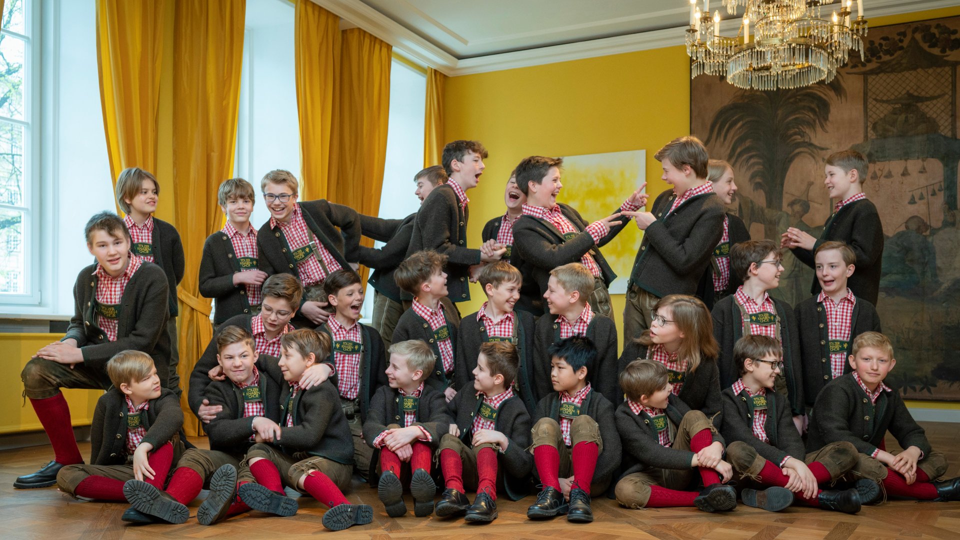 Kindergruppe in Tracht sitzt und spielt zusammen in einem hellen Raum mit gelben Vorhängen und Kronleuchter