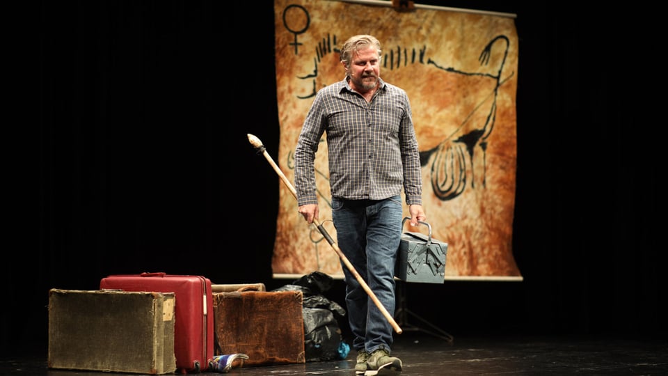 Foto für das Event mit dem Künstler auf der Bühne, im Hintergrund stehen vintage Koffer und eine prähistorische Felsmalerei