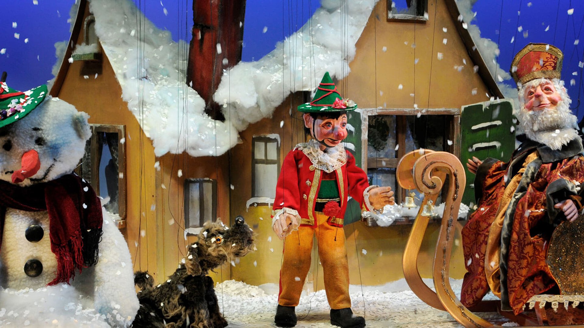 Winterliche Puppenszene mit Marionetten vor Häusern im Schnee; Figuren mit traditionellen Kostümen sowie Schneemann und kleiner Hund auf einer Bühne mit fallenden Schneeflocken.