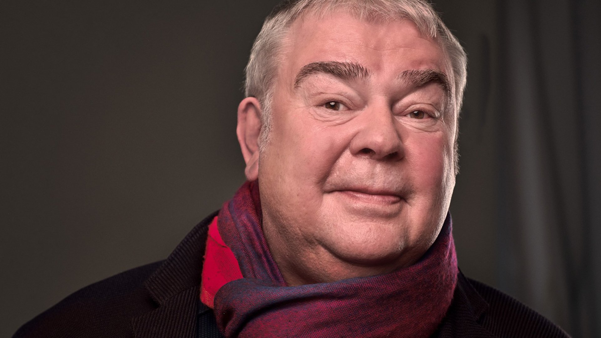 Portrait von Max Goldt mit rot-blauem Schal vor grauem Hintergrund