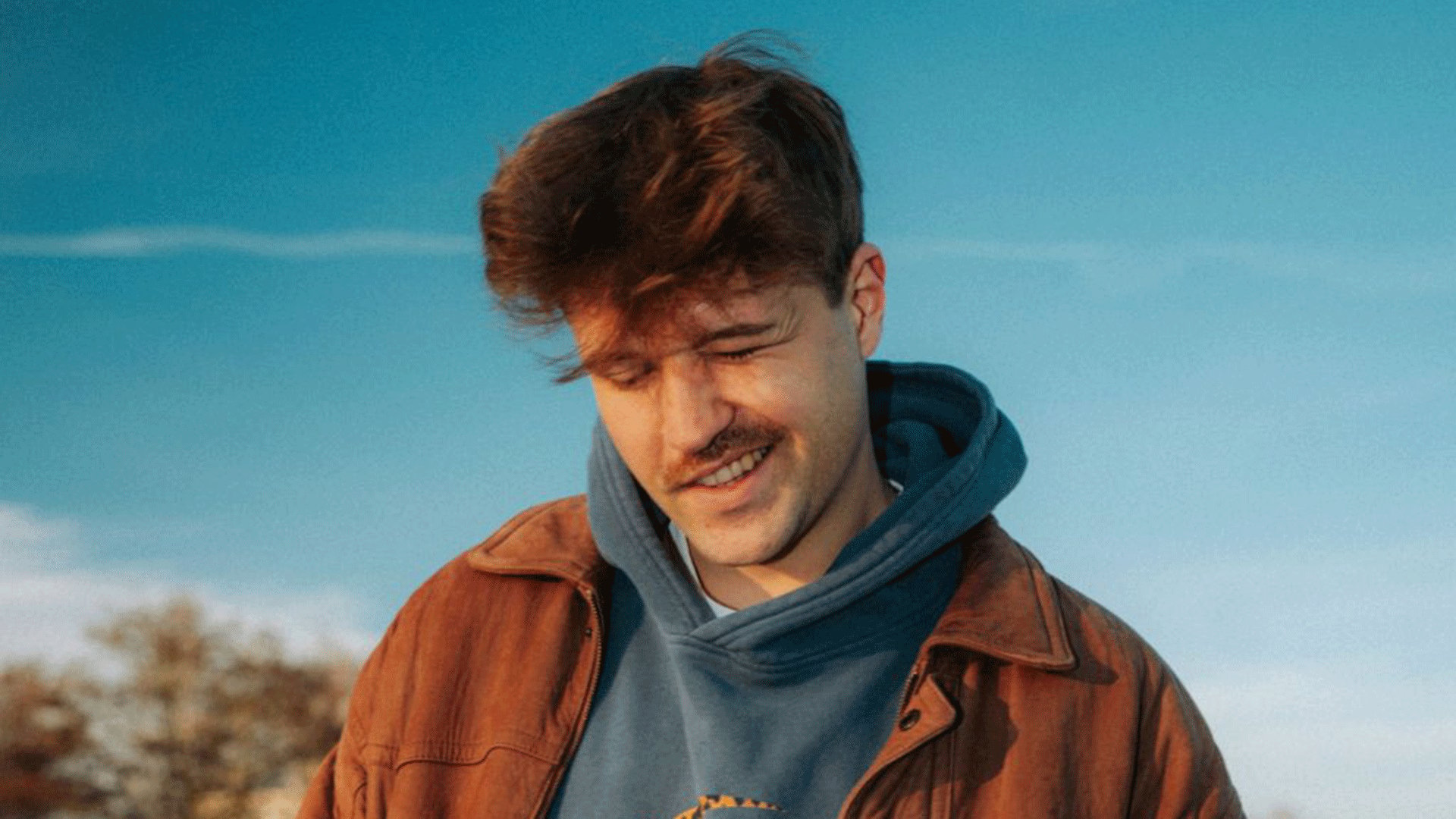 Ein Portrait vom Künstler mit blauem Hoodie und brauner Jacke an vor einem blauen Himmel
