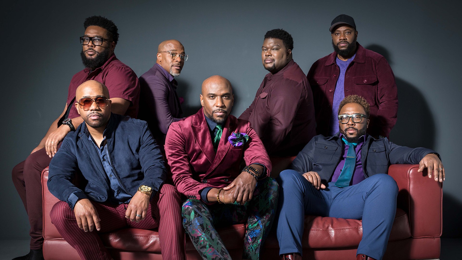 Sieben Herren in blauen und dunkelroten Anzügen auf einem dunkelrotem Ledersofa