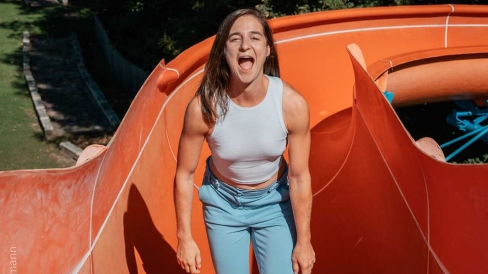 Eine Person mit weißem ärmellosem Top und hellblauer Hose steht in einer orangefarbenen Outdoor‑Rutsche; im Hintergrund sind Bäume und Grünflächen zu erkennen.