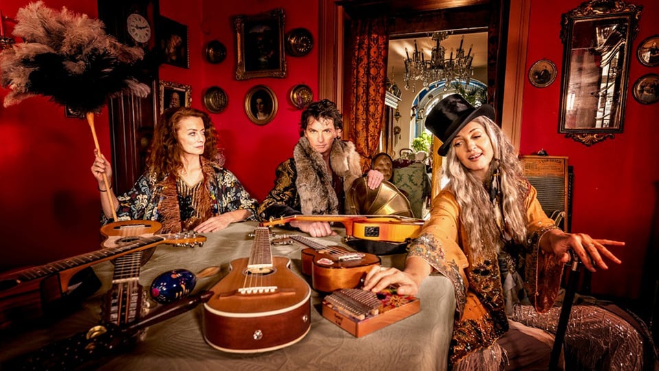 Hippie Kammerorchester: Drei Musikerinnen und Musiker sitzen in einem farbenreichen Raum im Vintage-Stil, umgeben von verschiedenen Instrumenten wie Gitarren und Ukulelen, warme Lichtstimmung.
