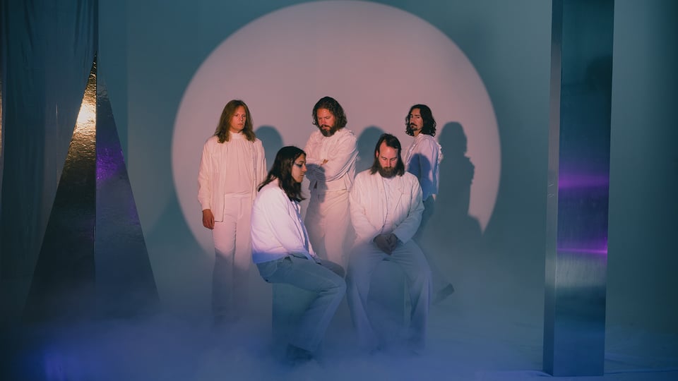 Die Band in weißer Kleidung vor einer Kulisse aus blauen, weißen und silbernen Formen