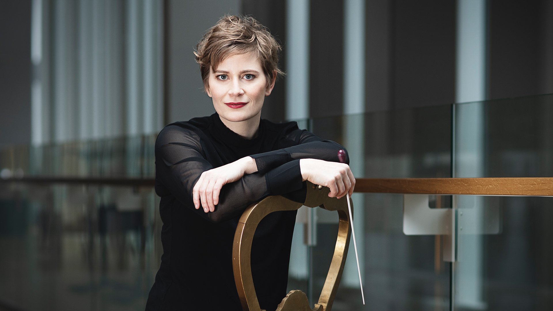 Anja Bihlmaier lehnt in schwarzer Kleidung an einem Geländer in einem modernen Innenraum und hält einen Taktstock in der Hand, im Hintergrund Glasflächen und architektonische Details