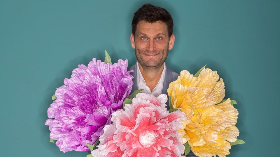 Foto für das Event „Christian Ehring – Versöhnung“ mit dem Künstler, umgeben von drei Blumen, vor einem blauen Hintergrund.