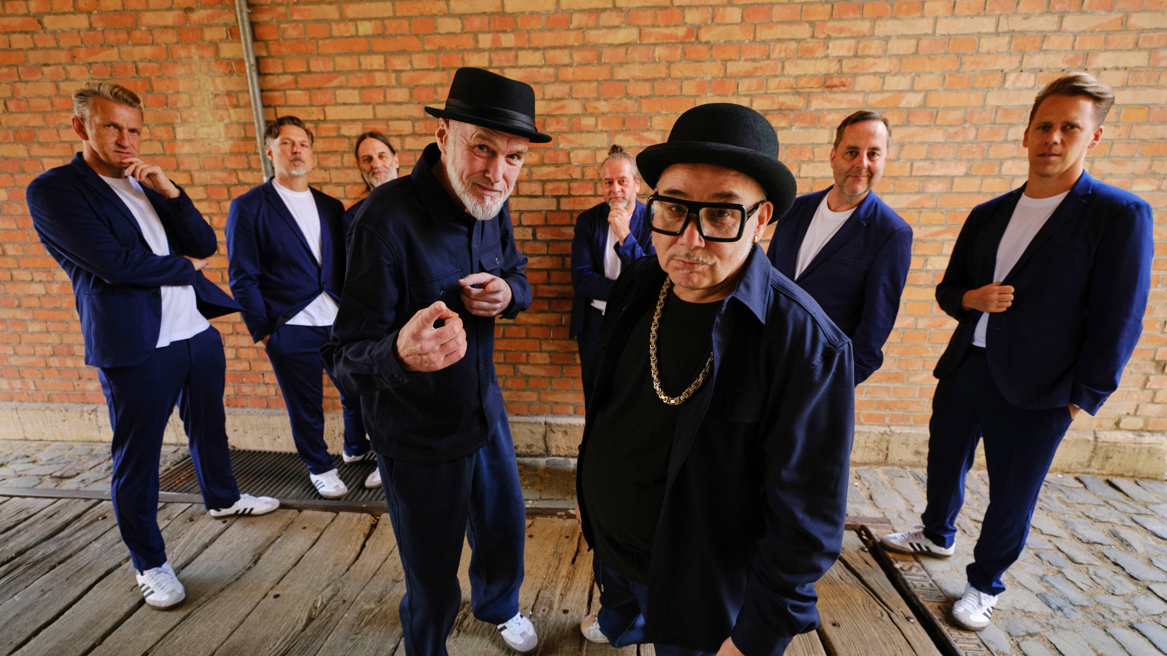 Bandfoto von der Jazzkantine vor einer Backsteinwand. Alle Bandmitglieder sind in blauen Anzügen und weißen Turnschuhen angezogen.