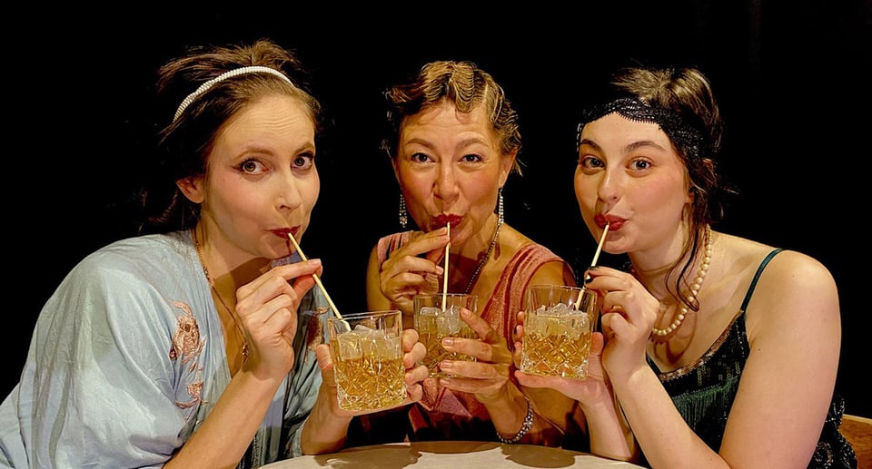 Drei Damen in Kleidung der 1920-er Jahre sitzen an Tisch, trinken aus Gläsern mit Strohhalmen