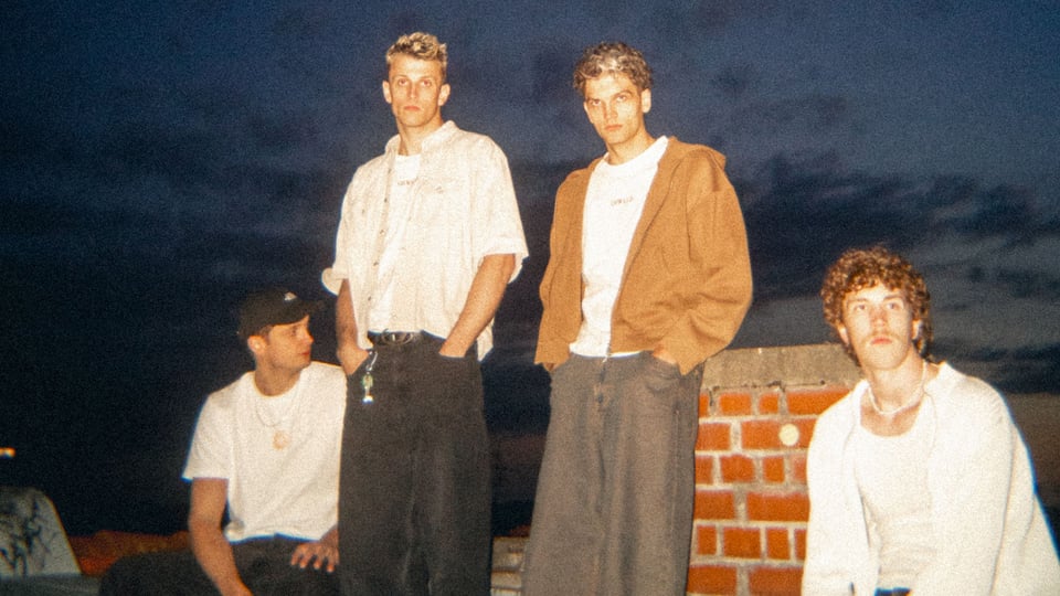 OSWALD Bandfoto: Vier junge Männer stehen und sitzen auf einer Mauer im Freien bei Abendstimmung, schauen in verschiedene Richtungen, dunkler Himmel im Hintergrund.