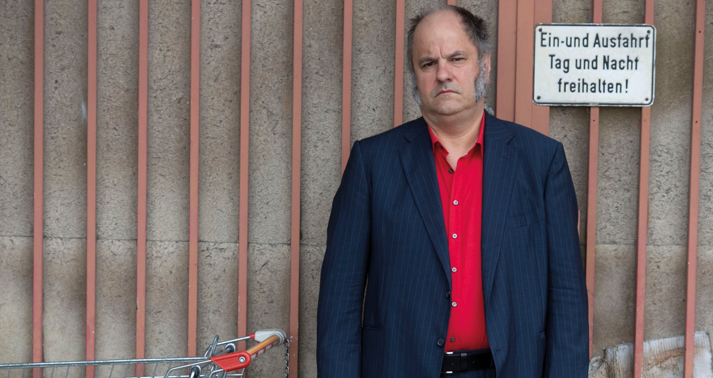Foto von „Matthias Egersdörfer“ in Anzugjacke und rotem Hemd. Im Hintergrund ein Tor und auf der linken Seite ein Einkaufswagen.