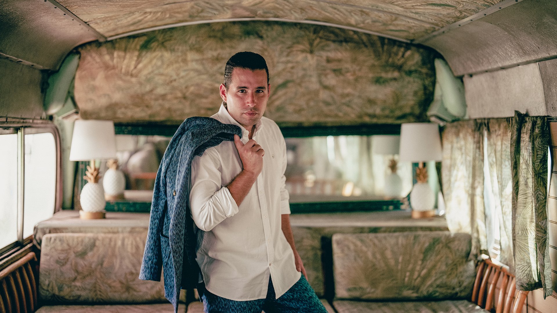 Alfredo Rodriguez in weißem Hemd und Jeans mit Sakko über der Schulter, im Hintergrund ein Raum mit beigen, opulenten Polstermöbeln und Wandausstattung