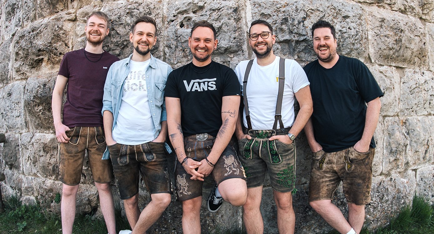Foto der Gruppe „d'Hundskrippln“ mit bayerischen Trachten, die an einer Steinmauer lehnen