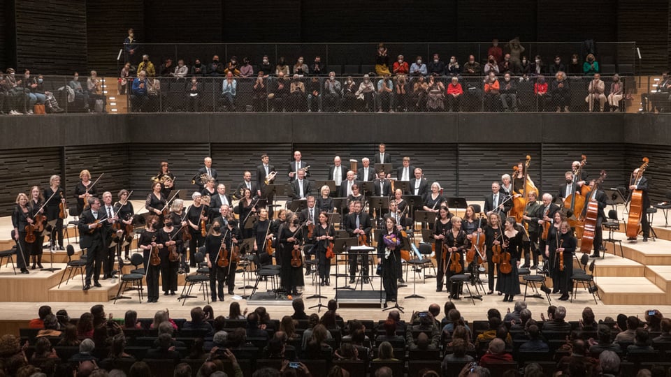 Bühnenfoto des Akademischen Sinfonieorchesters München mit großem Orchester auf der Bühne der Isarphilharmonie vor Publikum, Musikerinnen und Musiker stehen nach dem Konzert zwischen Streichern, Bläsern und Kontrabässen, während die Zuschauerränge im Hintergrund besetzt sind.