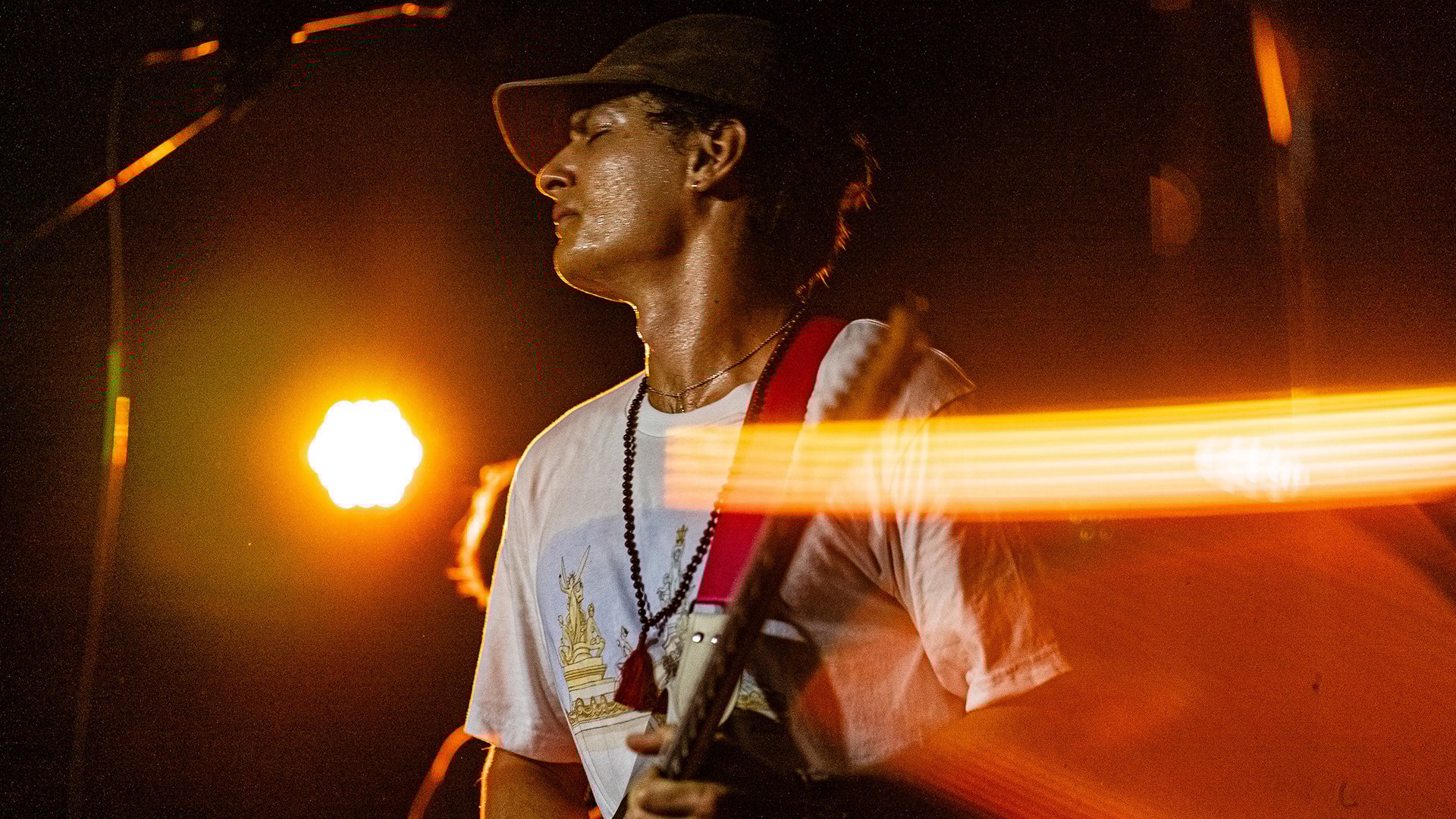 Der Künstler in weißem Shirt mit Kappe und Gitarre auf einer Bühne im Scheinwerferlicht
