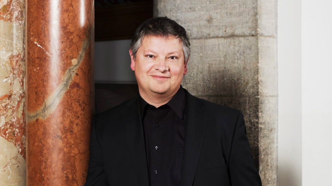 Porträtaufnahme einer Person in dunkler Kleidung, die seitlich an einer auffälligen marmorierten Säule lehnt; im Hintergrund sind eine Steinwand und ein Durchgang in einem Innenraum zu sehen, wodurch eine klassische architektonische Umgebung entsteht.