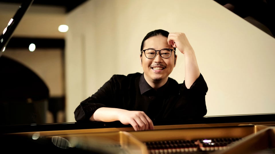 Porträt des Pianisten Kyohei Sorita an einem Flügel, halbnah aufgenommen, lehnt sich mit einem Arm auf das Instrument, trägt dunkle Kleidung und sitzt in einem Innenraum mit weicher Beleuchtung im Hintergrund.
