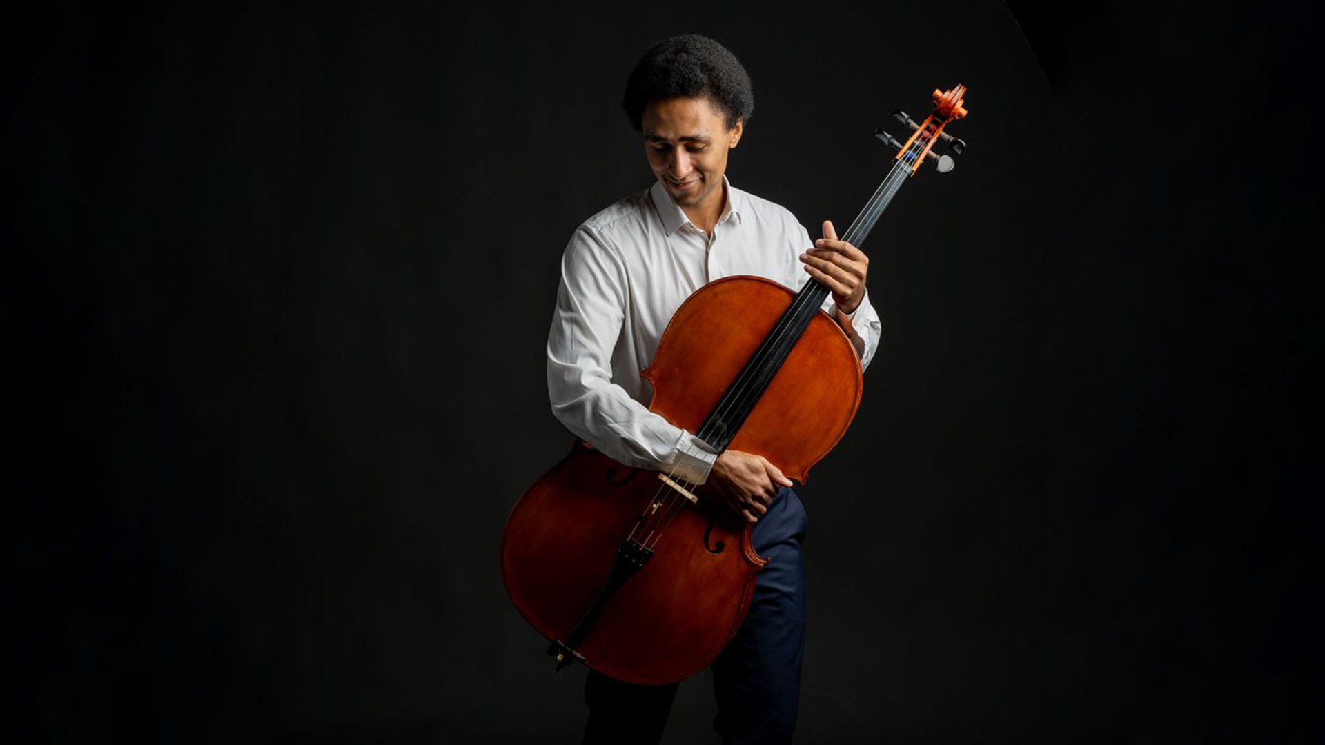 Studioaufnahme einer Person mit einem Violoncello: Die Person hält das braune Streichinstrument aufrecht und greift mit einer Hand den Hals, die andere ruht am Korpus; dunkler Hintergrund mit gezielter Lichtführung, Fokus auf Instrument, Haltung und Oberkörper, Darstellung eines klassischen Musikauftritts.