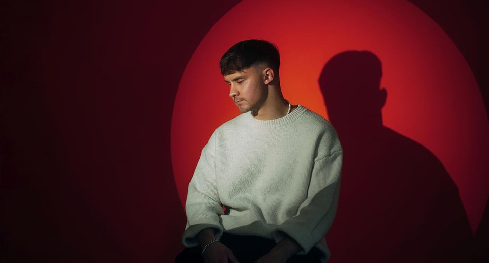 Foto des Künstlers mit einem weißen Pullover und rotem Licht im Hintergrund.