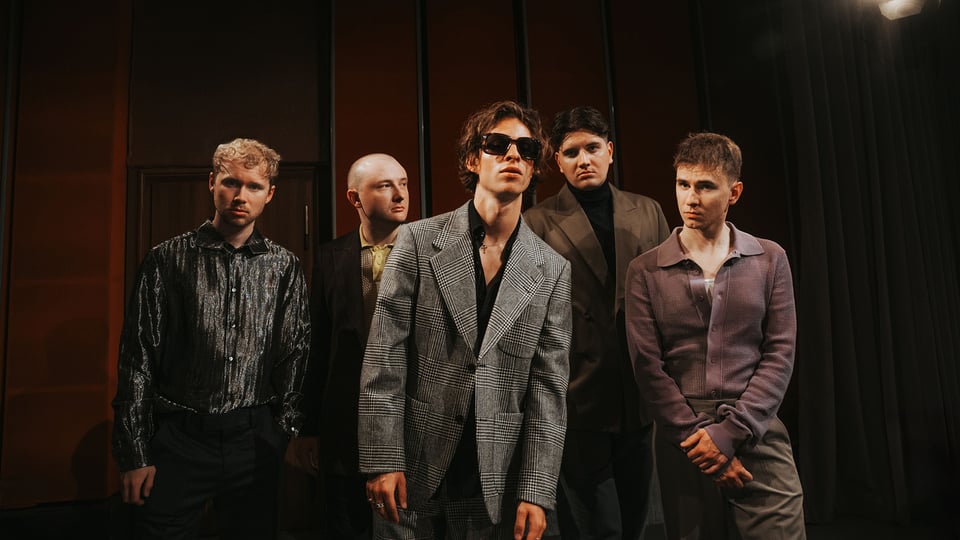 Die fünfköpfige Band steht vor einer dunkelrot vertäfelten Wand