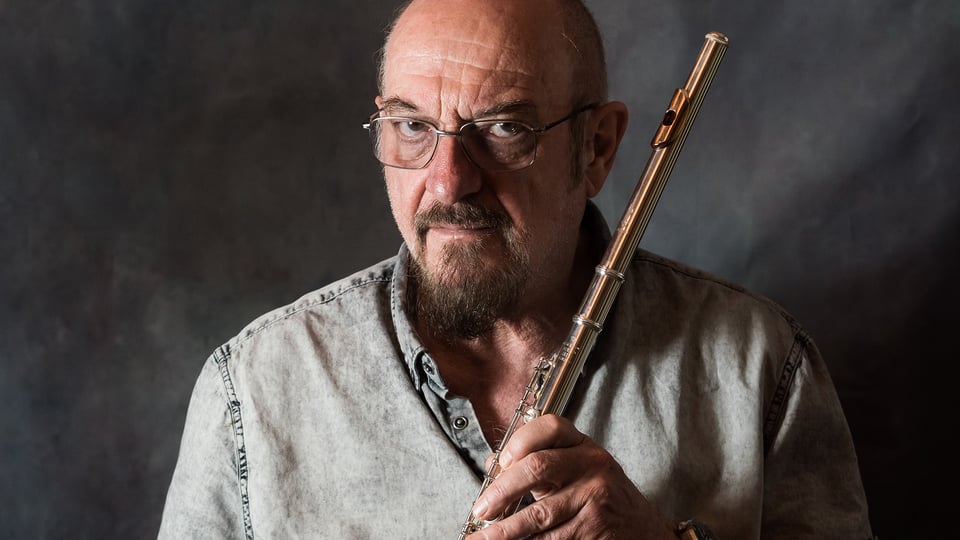 Promobild für das Event ‚Jethro Tull‘: Eine Person in einem hellen Jeanshemd hält eine Querflöte mit beiden Händen vor dem Oberkörper. Der Hintergrund besteht aus einer dunklen, strukturierten Fläche.