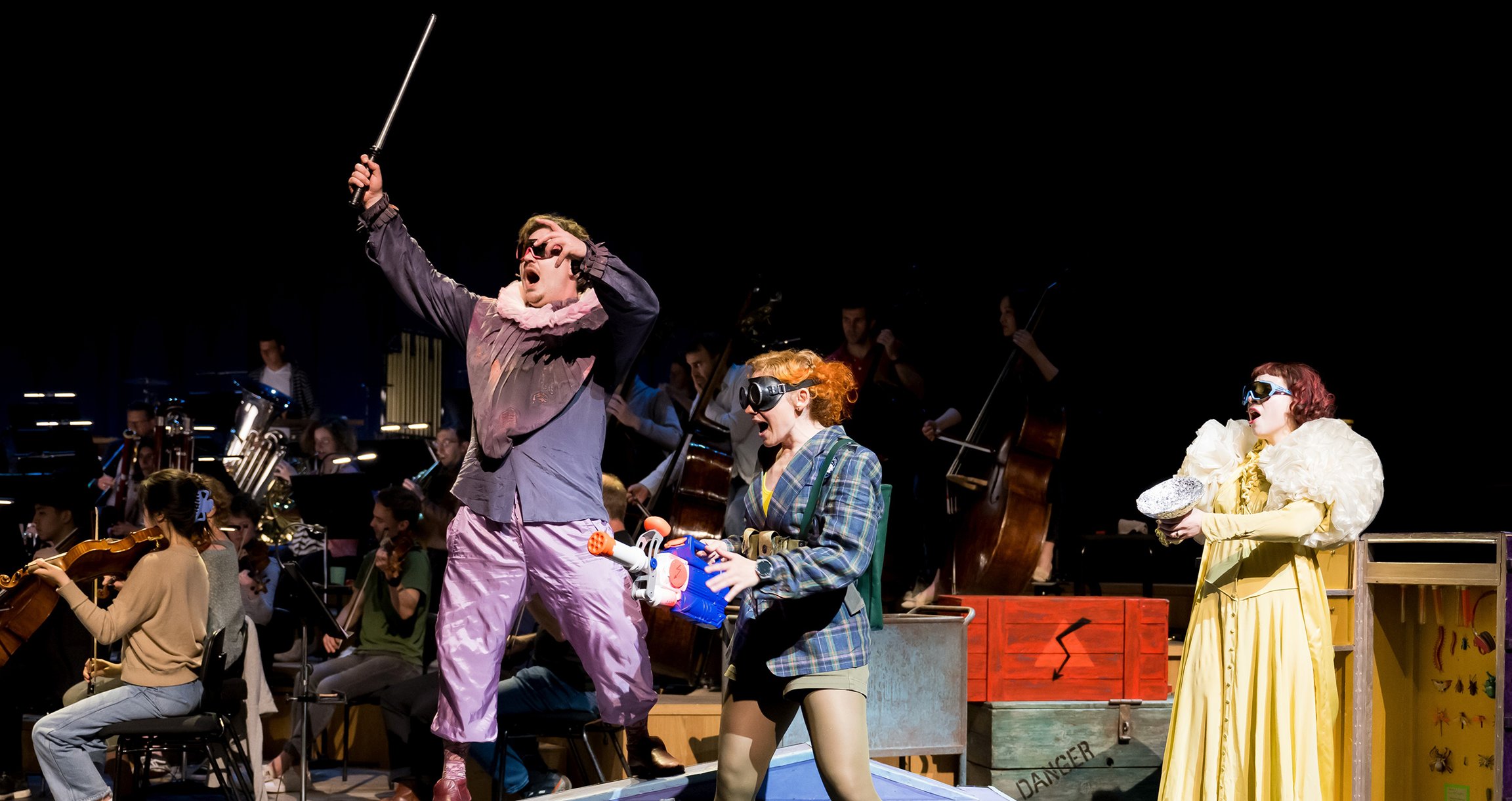 Ein Orchester spielt auf einer Bühne, während drei Schauspieler:innen in bunten Kostümen ein Theaterspiel für Kinder aufführen