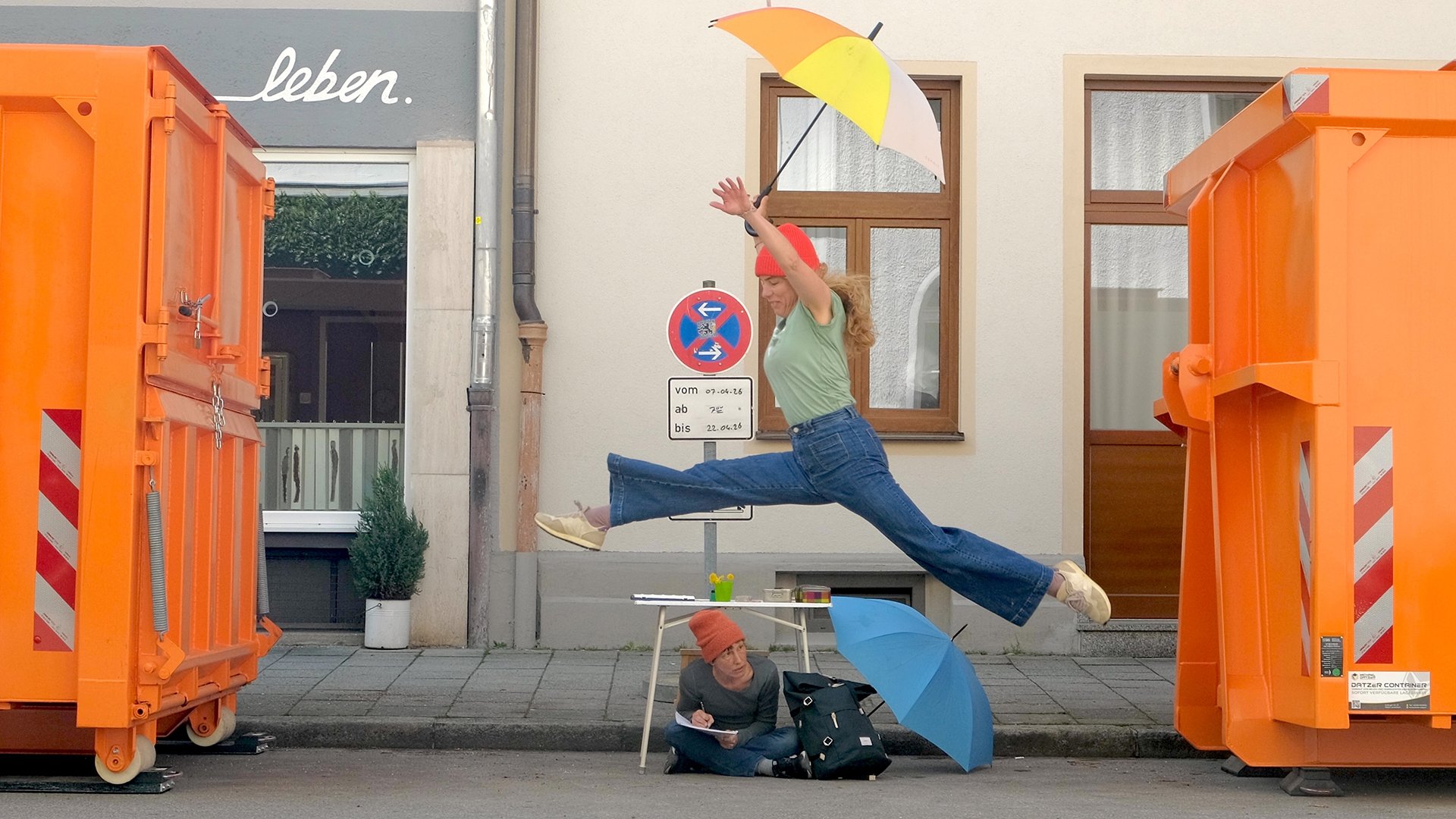 Zwei Personen zwischen Containern, eine springt mit Regenschirm, eine sitzt am Boden und schreibt vor Hausfassade.