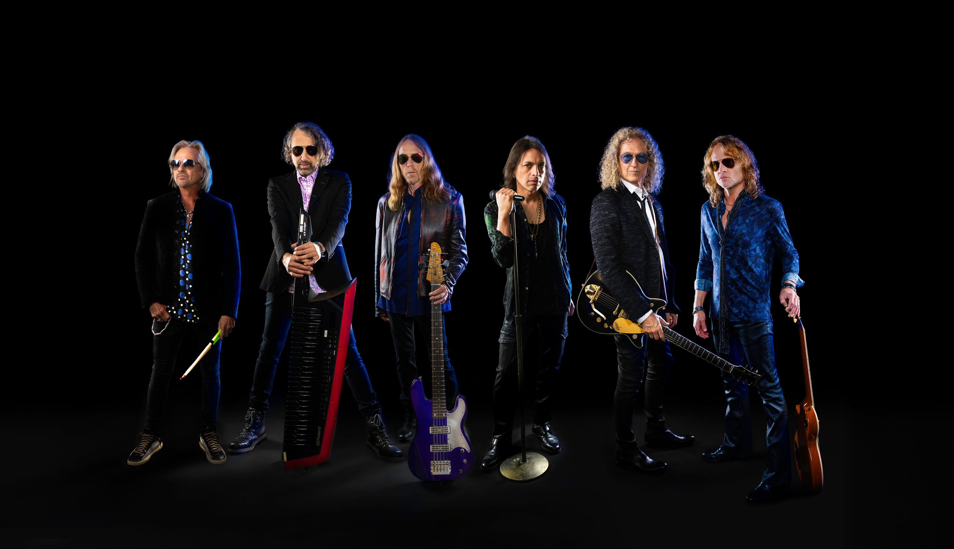 Die Band Foreigner in schwarzen und blauen Anzügen stehen mit instrumenten vor schwarzem hintergrund
