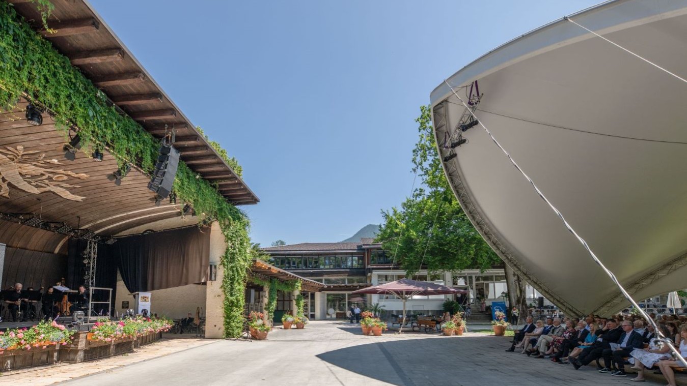 Freiluftkonzert auf einer Bühne in einem Innenhof mit Publikum unter einem großen Sonnensegel, Gebäude mit Pflanzenbewuchs und Sitzgelegenheiten im Hintergrund.