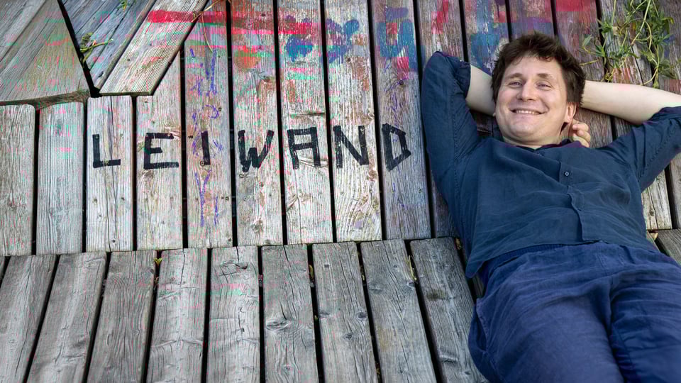 Eine Person, blau gekleidet, liegt entspannt auf hölzernen Planken, auf denen in schwarzer Schrift das Wort LEIWAND steht.