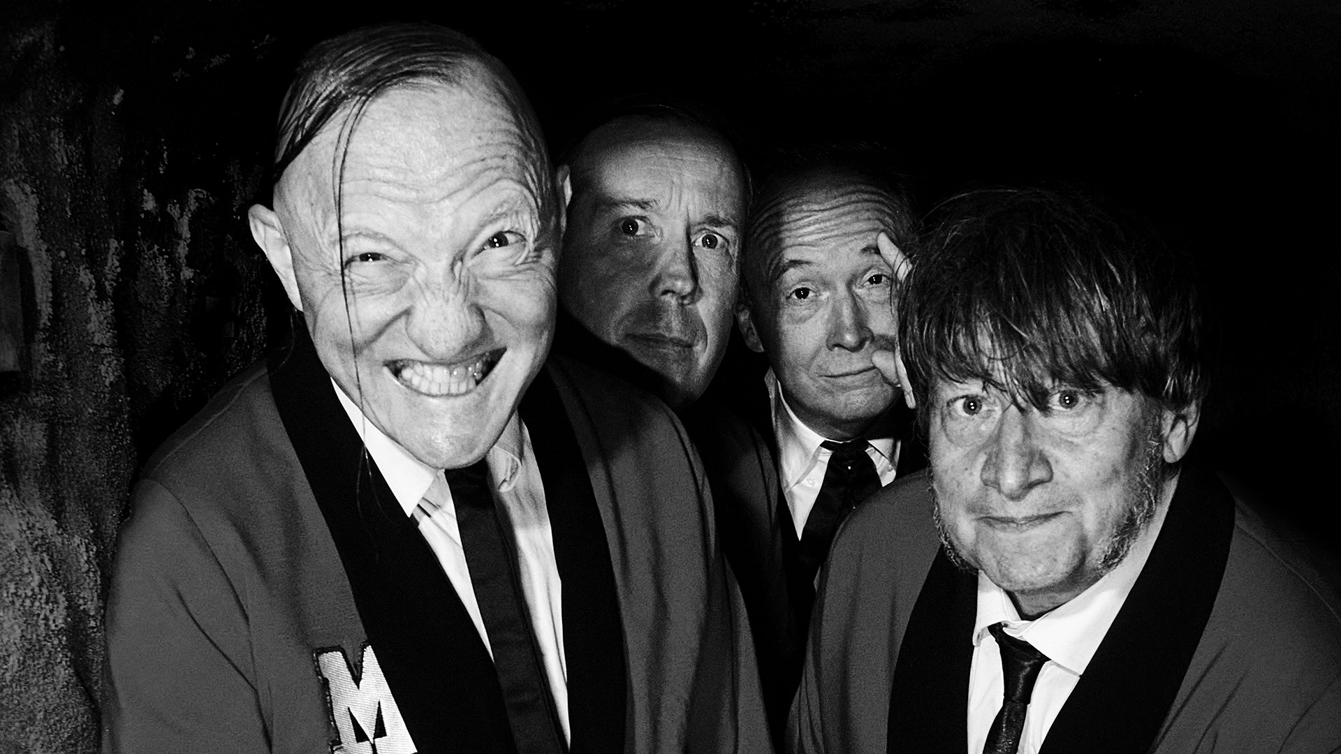 Bandfoto von The Monsters in Schwarz-Weiß. Vier Bandmitglieder in Anzügen und Krawatten stehen dicht vor dunklem Hintergrund und blicken mit markanten Posen direkt in die Kamera.
