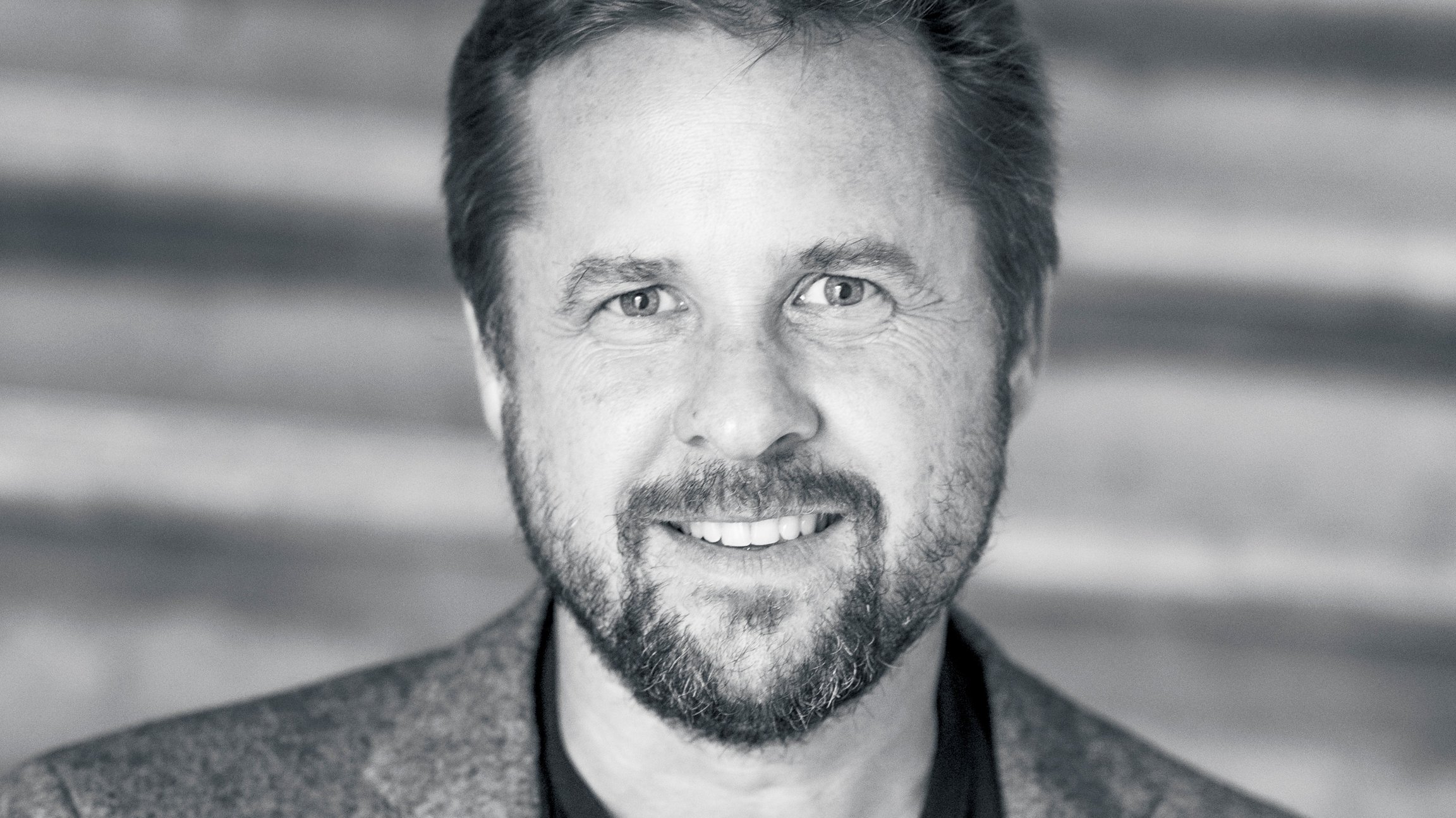 Porträt eines Mannes mit Bart und kurzem Haar in einem Sakko und T-Shirt, der lächelt und direkt in die Kamera blickt, unscharfer Hintergrund in Schwarz-Weiß.