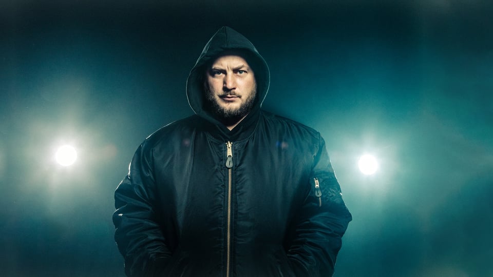 David Scheid steht mit Bomberjacke und Kapuze über dem Kopf vor einem dunkelnen Hintergrund.